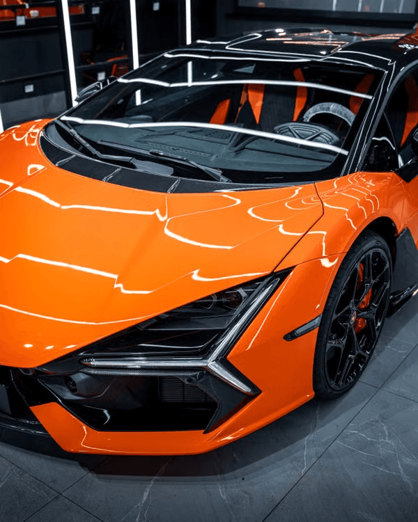Orange Lamborghini Revuelto front three-quarter under showroom lighting, with reflected light bars running across the bodywork